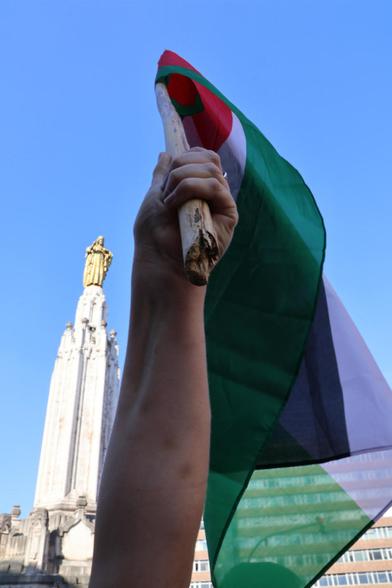En primer plano un brazo y puño sostienen una bandera palestina en alto. Al fondo, la figura del Sagrado Corazon de Jesús, sobre un pedestal. Es la plaza de dicho nombre en Bilbao.