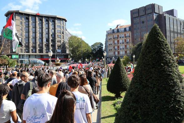 Multitud de gente con banderas palestinas, marchan en el lado izquierdo de la foto. A su derecha una zona verde con pequeños pinos y al fondo más árboles entre edificios altos. Es la Plaza Moyua de Bilbao
