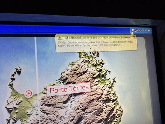 The image shows a computer screen displaying a detailed, topographical map of a landmass. The map is primarily green and brown, indicating vegetation and terrain, and appears to be a 3D rendering. A red marker with a white circle is positioned on the lower left side of the map. The name “Porto Torres” is written in red text near the marker. A yellow pop-up window is visible in the upper right corner of the screen with the following text: “Auf dem Desktop befinden sich nicht verwendete Dateien. Mit dem Desktop bereinigt kann der Desktop bereinigt werden. Klicken Sie auf dieses Symbol, um den Assistenten zu starten.” The computer's taskbar and clock are visible at the top edge of the screen, displaying a time of 13:48.