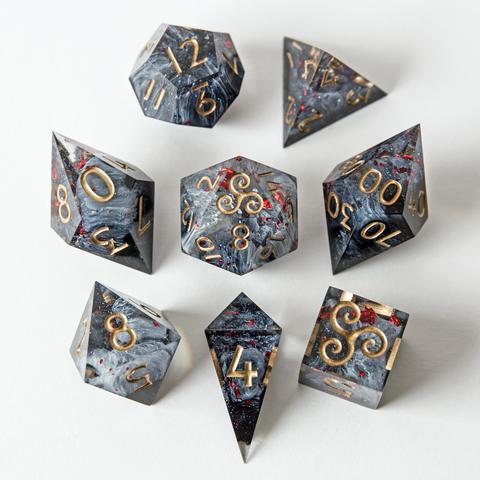 Ein Würfelset in Schwarz, Weiß und Grau mit rotem Blattgold im Inneren und Ziffern in Goldbronze. Das Set besteht aus einem D4-Shard, einer D4-Pyramide, einem D6, D8, D10, D100, D12 und D20.
Es liegt auf weißem Untergrund, jeweils mit der höchsten Seite nach oben. Beim D6 und D20 zeigt die höchste Seite anstelle einer Zahl eine Triskele. Die Würfel sind im Bild rund um den D20 angeordnet.