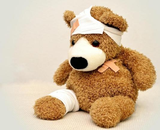 A close-up photograph of a fluffy, brown teddy bear sitting up. The bear is injured and wrapped in first aid bandages. A wide white bandage is wrapped around its head, secured with a couple of tan-colored adhesive bandages (band-aids). Another white bandage is wrapped around its lower left leg. A few more adhesive bandages are placed on its chest and arms. The bear has dark eyes, a black nose, and is set against a plain, light background.