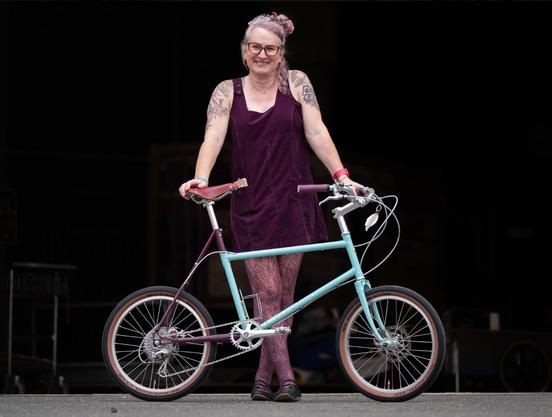 Elodie is standing behind a mint and purple mini velo with silver components
She is wearing a purple ish cord pinafore dress and matching patterned tights
Flower tattoos are visible across her shoulders and upper arms