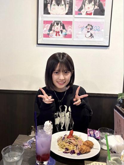 A young person is sitting at a restaurant table smiling and posing with a double peace sign. In front of them is a plate of pasta with fried chicken and a red sausage, along with a purple drink topped with whipped cream. Behind them on the wall are framed anime illustrations featuring a cheerful character with black hair in twin tails.