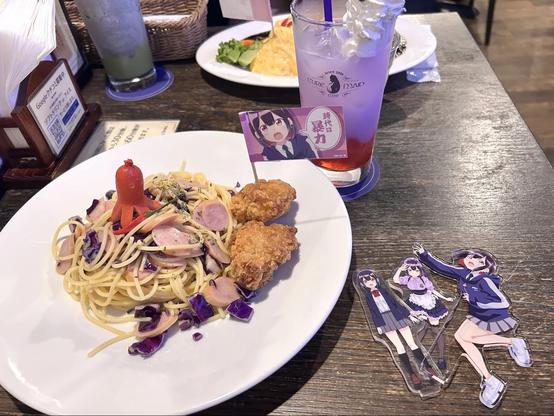 A close-up photo shows a themed meal featuring spaghetti with sausage, a small octopus-shaped sausage on top, and two pieces of fried chicken. Next to it is a purple drink with whipped cream, labeled “Cure Maid Café,” and a small flag with an anime character. On the table are acrylic standees of the same character in different outfits, matching the café’s anime theme.
