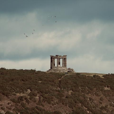 A neo-classical octagonal war memorial that resembles a ruined temple, seen from afar. A flock of birds fly above it.