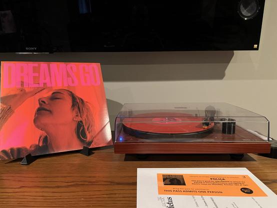 On the left standing up is the album sleeve for the album Dreams Go by Polica. It features the Channy's, the lead singer, head with water dripping down her face and neck. On the right my record player spins a red record.  In front of it lays the pass to see them live at the local record store tonight.