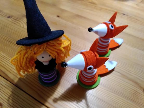 Im Vordergrund: Figuren aus Filz Scheiben, links eine Hexe mit vielen orangenen Haaren aus Wolle und einem schwarzen Filzhut. Rechts zwei Füchse aus orangenen und weißen Filz Scheiben, die Schnauze aus einem weißen Holzkegel mit schwarzer Holzperle an der Spitze. 
Im Hintergrund: Tischplatte aus Holz mit deutlich sichtbaren Schatten der Figuren.