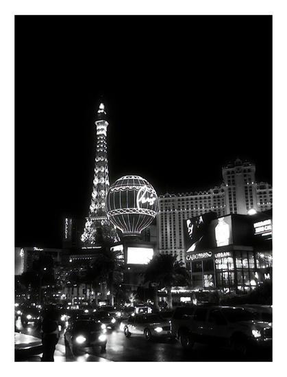 Black-and-white view of a bustling Las Vegas street at night, featuring illuminated Eiffel Tower replica and vibrant neon signs, evoking excitement.
