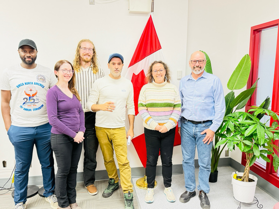 Four smiling people and one confused looking dude are standing in an office in front of a Canadian flag and some sort of broad leaf plant. It looks like they had a nice time chit chatting which indeed they did.