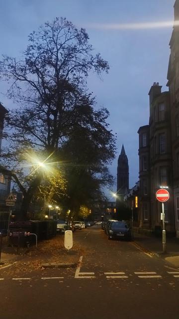 Looking past an illuminated "no entry" sign, down a leaf strewn street at dusk, tall, Victorian tenements, trees and a church spire silhouetted against the evening sky