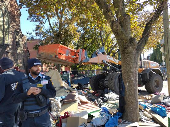 Photo of large machinery, nicknamed "the claw", demolishing encampment residents homes in front of St Stephens church, with 2 security guards in the foreground.