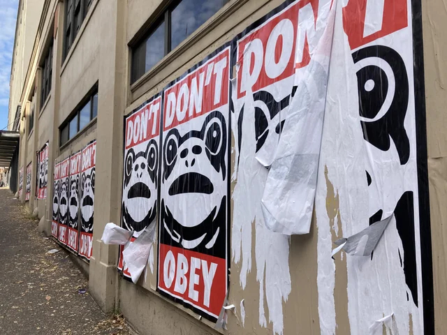 A photo of a building with a long row of identical large posters on the wall. The posters say "DON'T OBEY" with a drawing of the Portland inflatable frog's face.