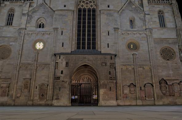 Das Foto zeigt das sogenannte Riesentor, den prächtigen Haupteingang des Stephansdoms in Wien, bei Nacht. Die Fassade des gotischen Bauwerks ist mit Steinreliefs und fein gearbeiteten Details verziert. Über dem mittigen Spitzbogenportal erhebt sich ein großes gotisches Fenster. Die Szene ist ruhig, die Beleuchtung betont die architektonischen Strukturen und lässt die imposante Größe des Portals und der umgebenden Mauern deutlich werden.