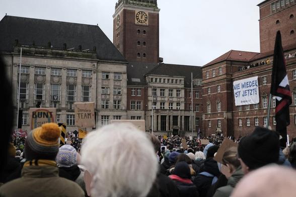 Eine große Menschemenge steht auf dem Kieler Rathausplatz. Am Opernhaus rechts hängt ein Banner mit der Aufschrift "Nie wieder ist jetzt". Ein Plakat mit einem Regenbogen und eins mit der Aufschrift "Demokratie Team" sind zu erkennen.