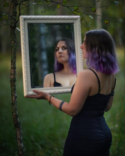 a woman holding a mirror in front of a pond looking at the camera from the reflection