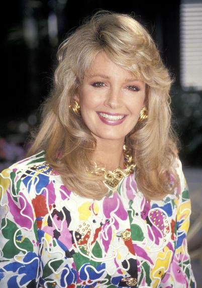 A professionally shot 1991 portrait of Deidre Hall at the NBC Summer TCA Press Tour. She stands smiling directly at the camera, wearing a vibrant multicoloured abstract floral jacket with white piping over a white skirt. Her shoulder-length blonde hair is softly layered and styled with volume. She accessorizes with bold gold jewelry, including large earrings and a statement brooch, adding to the dramatic flair of the outfit. The background is softly blurred in natural tones, giving the image a warm, elegant feel. Her expression is bright and confident, capturing the glamour, confidence, and iconic presence she brought to her role as Marlena Evans on Days of Our Lives.