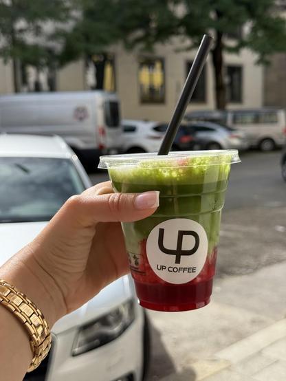 A hand holding a green and red smoothie with a black straw, labeled "UP COFFEE," in front of a city street.