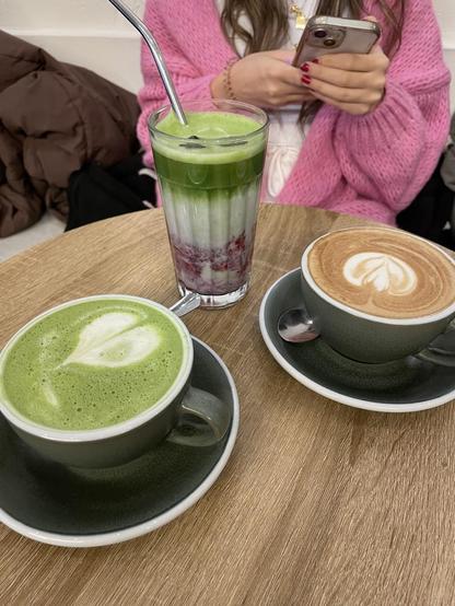 A table with a matcha latte, a regular latte, and a matcha drink with berries, next to a person on their phone.