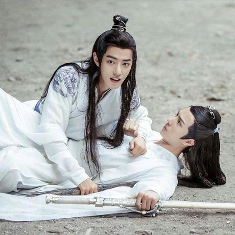 Teenage Wei Wuxian and Lan Wangji are sprawled out across the ground, Lan Wangji on his back with Bichen clutched in his left hand where it is outstretched on the ground in the direction of the viewer and Wei Wuxian across him, stomach to stomach. Wei Wuxian is leaning on his left arm, which is across Lan Wangji's upper chest and bound to Lan Wangji's right arm with the sacred Lan forehead ribbon, while Suibian is in his right hand where it rests on the ground between Lan Wangji's torso and long sleeve. Wei Wuxian is looking in the direction of the viewer at an off screen target with his mouth open just enough that his upper teeth are visible while Lan Wangji has raised his head a few inches off the ground to glare at Wei Wuxian.