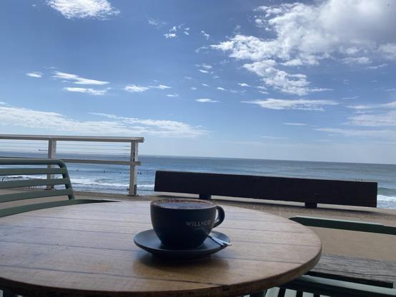 Cappuccino in a black cup and saucer on a low wooden table overlooking the southern end of Merewether Beach. Blue sky with patches of white cloud. Blue water with a fringe of white.