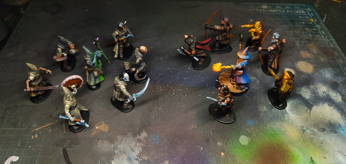 Deux bandes de figurines peintes pour Frostgrave disposées face à face sur un plan de travail. À gauche, des mages et soldats encapuchonnés aux tons gris et verdâtres. À droite, un groupe plus coloré, dominé par un mage en robe bleue tenant une flamme magique.