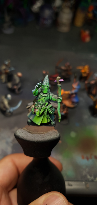 Gros plan sur un mage encapuchonné peint en dégradé de vert et gris, tenant un bâton orné de cristaux violets. Son visage est caché, seule une main bleuâtre luit d’énergie occulte.