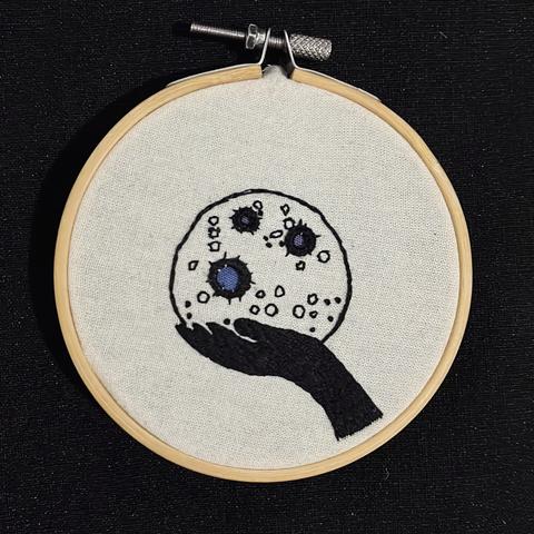 Una obra bordada que presenta una mano negra que sostiene una luna, con diversas texturas y colores, sobre un fondo negro. La pieza está enmarcada en un bastidor de bordado de madera.