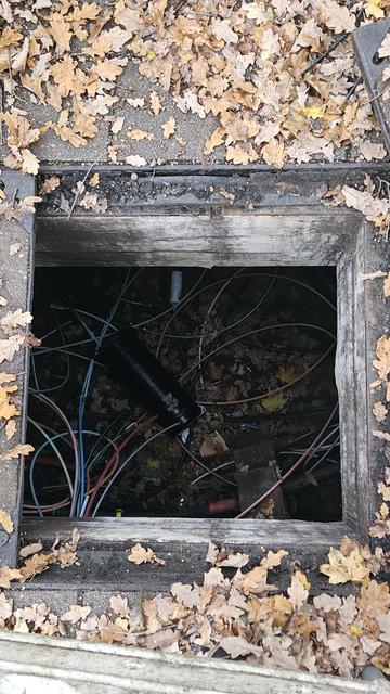 Photo in einen länger offenstehenden Kabelschacht mit chaotisch rumliegenden Glasfaserkabel. Es sammelt sich Laub auf dem Gehweg und unten im Schacht.