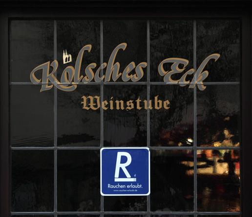 das bild zeigt ein fenster mit dunklem rahmen und mehreren feldern, die durch stege getrennt sind. auf dem fenster befindet sich in geschwungener schrift der name "kölsches eck" und darunter "weinstube", wobei über dem "o" von "kölsches" eine stilisierte abbildung des kölner doms zu sehen ist. unter der schrift befindet sich ein blaues schild mit einem großen weißen "r" und dem text "rauchen erlaubt" sowie einer webadresse. das fensterglas ist leicht strukturiert. es reflektiert etwas und es ist kneipenlicht im inneren des raumes sichtbar.