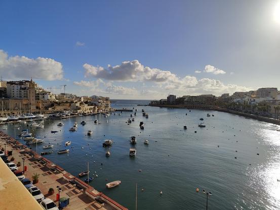 Building sit either side of a small harbour filled with small boats. The ocean is visible beyond the harbour mouth. A few puffy clouds today against a blue sky.