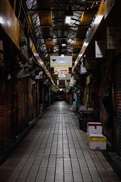 Empty covered night market with tiled walkway flanked by shuttered vendor stalls, fluorescent lights illuminating yellow support beams and hanging signage under corrugated metal roof