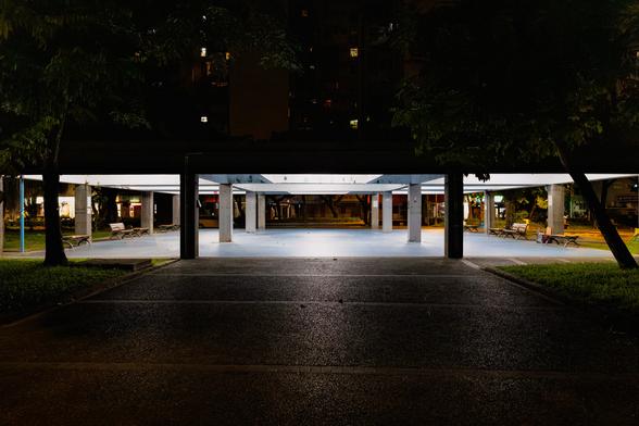 Illuminated concrete pavilion with flat roof and square pillars in a Taipei park at night, park benches underneath, framed by silhouetted trees with apartment building lights visible in background