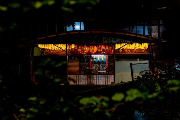 Taipei Nantian Temple at night with ornate curved roof, rows of glowing yellow and red paper lanterns beneath decorative eaves, metal gate at entrance, viewed through dark foliage
