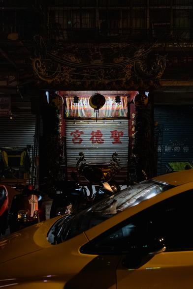 Shengde Temple entrance at night with closed white metal shutters displaying red Chinese characters 聖德宮, ornate gold-painted archway above, yellow taxi and motorcycles parked in front