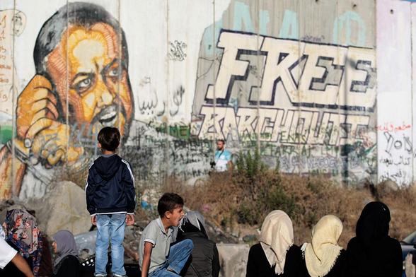 Niños y adultos palestinos esperan junto al muro israelí ilegal para cruzar en el puesto de control de Qalandia, con una pintada del preso Marwan Barghouti al fondo, en 2015. (NurPhoto via Getty Images)