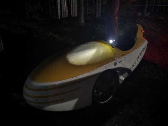 Keltavalkoinen kinneri eli kolmipyöräinen katettu nojapyörä hämärässä. Pyörän etuvalo valaisee kinnerin kuomua, jossa on valkoista huurretta. Vieressä on mäntyjä.