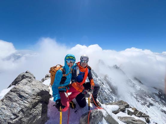 two people at the top of a high mountain peak, wearing helmets and crampons. clouds and blue sky in the background