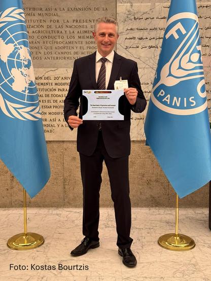 Prof. Dr. Marc F. Schetelig steht zwischen zwei blauen Fahnen mit dem weißen Logo der FAO und hält eine Urkunde in der Hand.