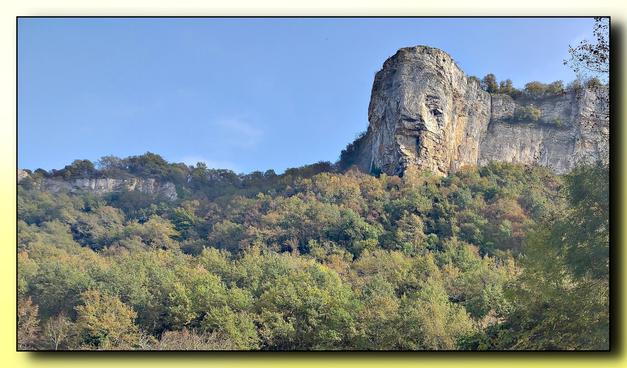 Au delà d'une pente boisée parée des premières couleurs d'automne s'élève une falaise calcaire illuminée par le soleil. Le ciele est bleu, sans nuage.