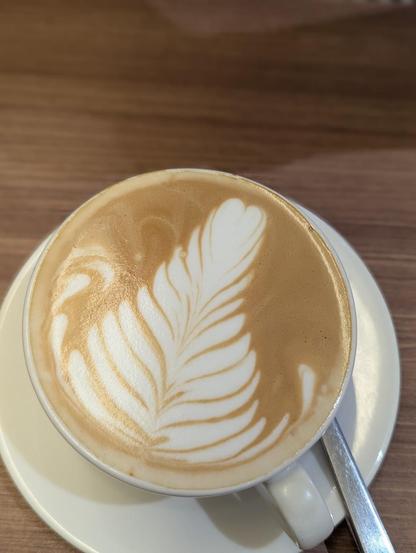A latte with a feather design in the foam, in a white cup and saucer, with a spoon on a wooden table.