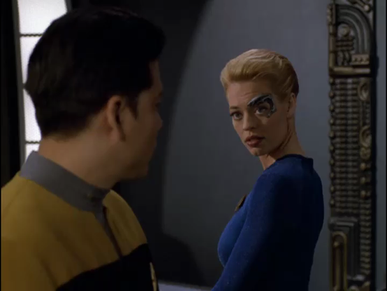La imagen muestra a dos personas en un interior oscuro, probablemente de una nave espacial.

En primer plano y de espaldas al espectador, se ve la cabeza y el hombro de un hombre con cabello oscuro corto. Viste un uniforme de Starfleet de color amarillo y negro con un cuello gris.

Frente a él, y mirando hacia el espectador por encima de su hombro derecho, hay una mujer rubia con el cabello recogido. Lleva un distintivo implante cibernético plateado sobre su ojo izquierdo y parte de su sien. Viste un ajustado traje azul de manga larga y tiene una expresión seria o ligeramente sorprendida, con los labios entreabiertos.

El fondo es gris oscuro, con algunos detalles arquitectónicos metálicos en la pared derecha.