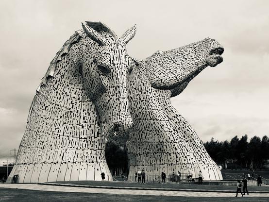 Two huge steel horse heads next to the M9 motorway in Scotland. Small people walk around them, taking pictures.