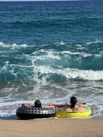 Two people sit on inflatable tubes at the edge of a beach, facing the ocean waves. One tube is black with a tire pattern, and the other is yellow with a pineapple design. The ocean is blue with white-capped waves.

Zwei Menschen sitzen auf aufblasbaren Röhren am Rande eines Strandes, mit Blick auf die Wellen des Ozeans. Ein Schlauch ist schwarz mit einem Reifenmuster und der andere ist gelb mit einem Ananas-Design. Der Ozean ist blau mit weißen Wellen.