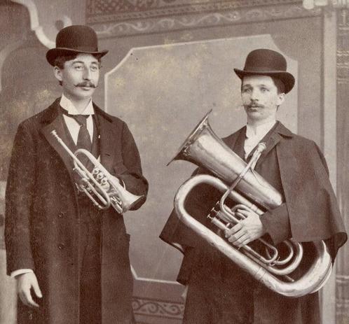 A vintage sepia tone photo of two mustachioed and be-hatted men holding a tuba and a trumpet.