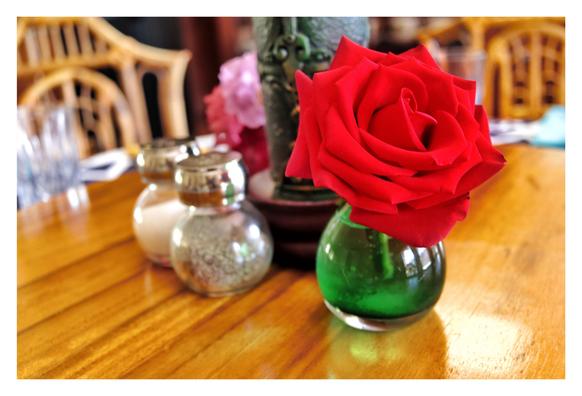 Photo of a perfect red rose with velvety petals sitting in a small, round, green vase, on a Lazy Susan with a glossy sheen over its wood grain. Behind and to the left of the rose are salt and pepper shakers and a large green decorative candle with a molded surface pattern. Further back are drinking glasses and pink and purple blossoms in vases, and at the edge of the dining table are rattan or bamboo chairs. Everything is slightly out of focus except for the rose.
