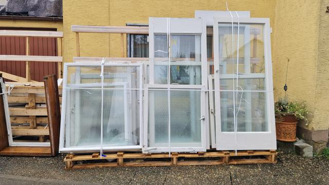 Ausgebaute ganze Fenster mit weißen Holzrahmen hochkant auf Palette Festlegungen stehn vor Hauswand.