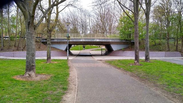 A pale blue/ grey bridge over a path running through a park. The path dips gently down to get under the bridge. there are mature trees all around.