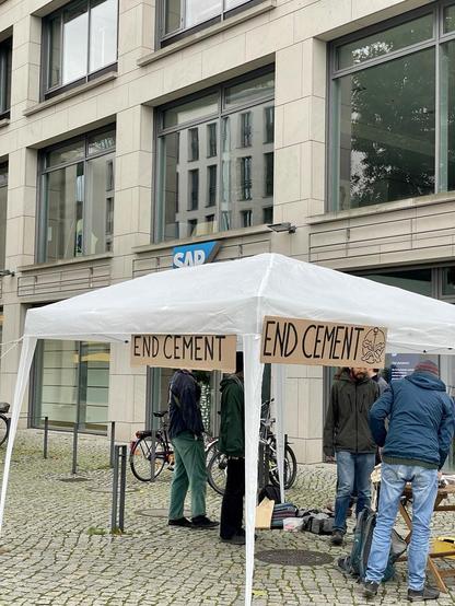 Mit End Cement beschrifteter Pavillon vor einem Haus mit SAP Beschriftung