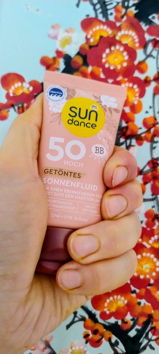 Fingers of a light-skinned left hand around a tub of sun screen which says "50 hoch" and "getöntes sonnenfluid" on it