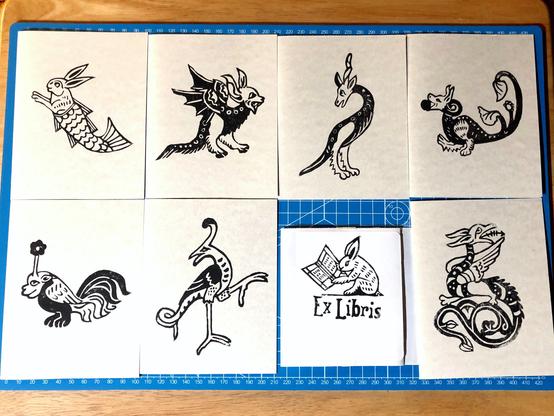 seven block printed cards on white card stock, and one 3/75x4 label laid out on a blue cutting mat.
the card prints are of a merbunny, a small lizard creature with feathery wings and a devils face, a tall two legged creature with a long tail, furry legs and a horses head and unicorn horn, a small lizard like creature with two furry legs, a long split tail with leaved at the end, and a dog's head carrying a ball in it's mouth, a small creature with two pawa, a rooster's tail, small wings and a face like a smug bulldog with a flower growing out of it's head, a tall stork like creature with a lizards tail with one leg lifted as if to grab something, a small wyvern like creature with an arrow tongue, feathery wings standing on it's very long, split and curling tail tipped in filigree leaves, and the bookplate is of a small bunny reading an open manuscript with the words Ex Libris underneath.